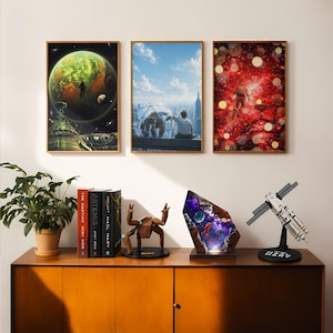 May include: Three framed space-themed posters hang above a wooden cabinet featuring a potted plant, three books, a brown geometric creature figurine, a glowing resin lamp with an astronaut scene, and a white and black satellite model on a stand.