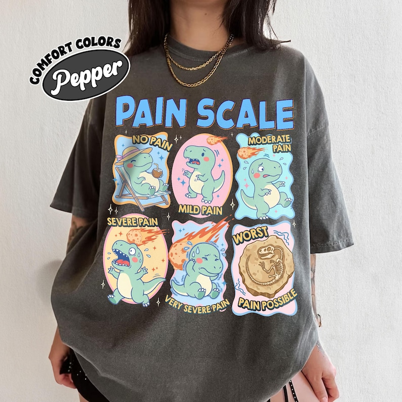 Op de afbeelding: Donkergrijs t-shirt met een kleurrijke afbeelding van een pijnscore met cartoon dinosaurussen. De tekst op het shirt luidt "PAIN SCALE" en bevat niveaus van "No Pain" tot "Pain Possible". Het shirt heeft ook de tekst "COMFORT COLORS Pepper".