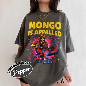 Può includere: T-shirt grigio scuro con la scritta gialla "MONGO IS APPALLED". La grafica presenta un gatto con occhiali da sole e una corona in sella a una creatura colorata simile a un dinosauro. La maglietta ha un'etichetta Comfort Colors Pepper.