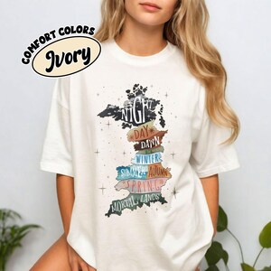 以下が含まれることがあります： アイボリーのTシャツ。イギリスの地図が水平なセクションに分割されたグラフィックが描かれており、Night、Day、Dawn、Winter、Summer、Autumn、Spring、Mortal Landsというテキストラベルと、小さな星のイラストが周囲に配置されています。