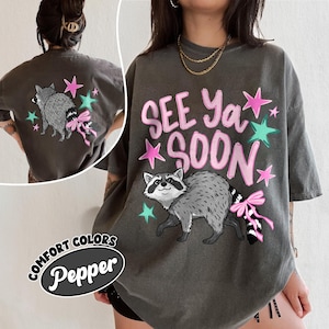Puede incluir: Camiseta gris oscuro con un gr&aacute;fico de mapache y el texto "SEE Ya SOON" en rosa. El mapache tiene un lazo rosa en la cola, con estrellas rosas y turquesas. La camiseta es Comfort Colors Pepper.