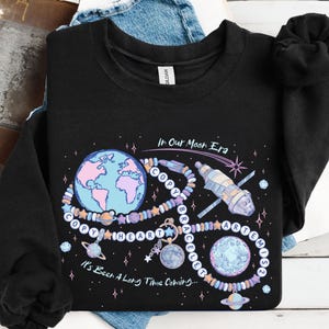 Puede incluir: Sudadera negra con un diseño gráfico colorido que presenta un planeta Tierra, una pulsera, una nave espacial y el texto "In Our Moon Era" y "It's Been A Long Time Coming...". El diseño incluye colores rosa, azul y morado.