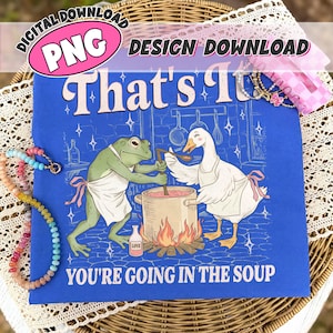 Op de afbeelding: Een koningsblauw t-shirt met een grafische illustratie van een groene kikker en een witte gans in schorten die samen koken. De tekst luidt That's It en You're Going In The Soup. Een klein flesje met het label Love staat bij de kookpot.