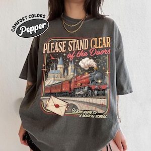 May include: A charcoal gray short-sleeve t-shirt featuring a graphic of a red steam locomotive traveling toward a castle. The text reads PLEASE STAND CLEAR of the Doors and WE'RE GOING TO A MAGICAL SCHOOL, with an envelope icon below.