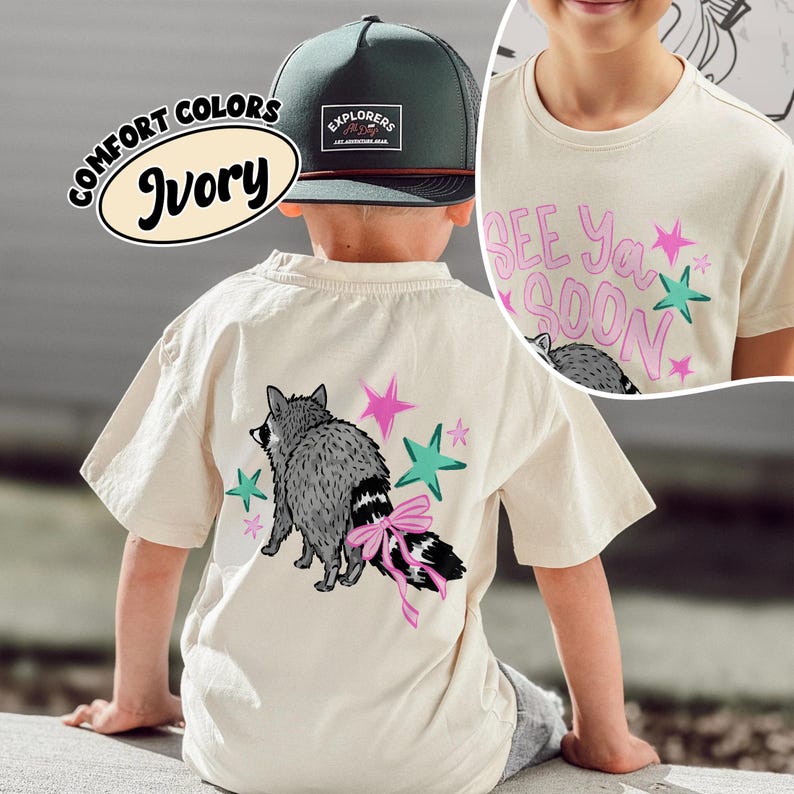 Puede incluir: Camiseta color marfil con un gr&aacute;fico de mapache y el texto "See Ya Soon". El mapache tiene un lazo rosa en la cola, con estrellas rosas y turquesas. Tambi&eacute;n se ve una gorra verde y negra con "Explorers". Color confort.