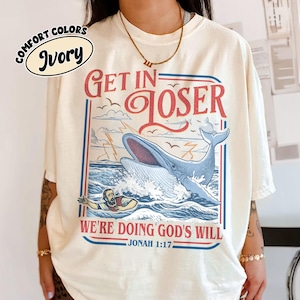 Op de afbeelding: Ivoor t-shirt met een afbeelding van een blauwe walvis, een persoon in het water en de tekst "Get in Loser, We're Doing God's Will". Het ontwerp bevat rode en blauwe accenten, vogels en bliksem. De tekst "Jonah 1:17" is ook opgenomen.