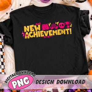 May include: A black t-shirt featuring the yellow text NEW ACHIEVEMENT! with a pink exclamation point. Above the text are small pink icons including patterned shorts, sunglasses, a skull, and a trophy, accented by yellow lightning bolt graphics.