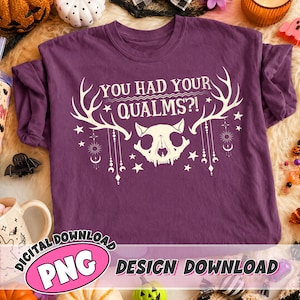 May include: A purple short-sleeve t-shirt featuring a white graphic print of an animal skull with large antlers. The text "YOU HAD YOUR QUALMS?!" is printed above the skull, surrounded by celestial symbols like stars, suns, and moons.