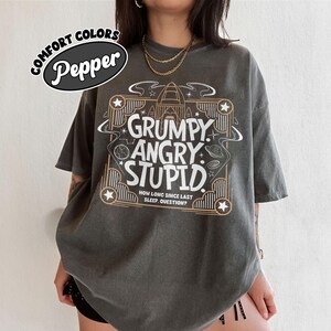 May include: A dark gray t-shirt with the words "GRUMPY, ANGRY, STUPID" in a decorative frame. The shirt also features the text "HOW LONG SINCE LAST SLEEP, QUESTION?" and the words "COMFORT COLORS" and "Pepper".