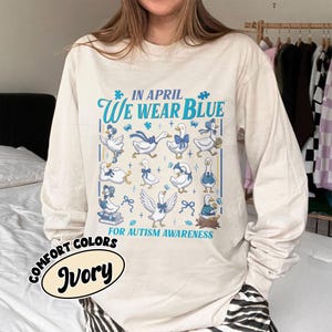 May include: Ivory long-sleeve shirt with the text "IN APRIL WE WEAR BLUE FOR AUTISM AWARENESS." The shirt features illustrations of geese with blue accents. The words "COMFORT COLORS" and "Ivory" are also printed on the shirt.