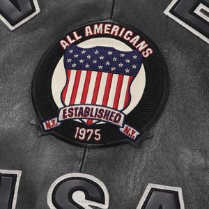 Puede incluir: Primer plano de una chaqueta de cuero negra con un parche circular. El parche presenta el texto "ALL AMERICANS" en rojo, un dise&ntilde;o de escudo con rayas rojas y blancas y estrellas azules, y "ESTABLISHED 1975 N.Y."