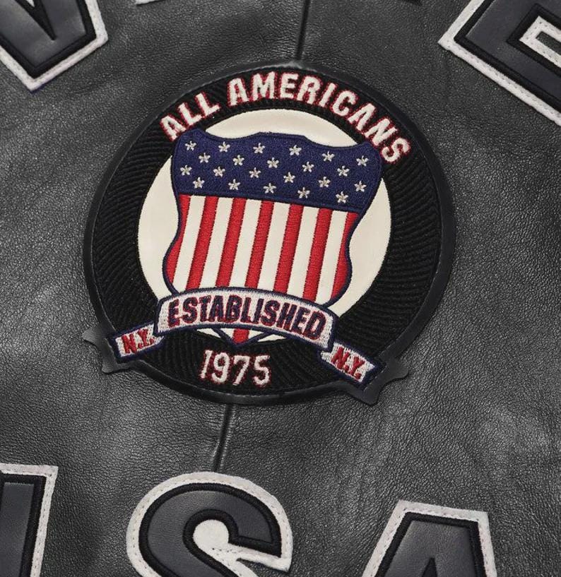Puede incluir: Primer plano de una chaqueta de cuero negra con un parche circular. El parche presenta el texto "ALL AMERICANS" en rojo, un dise&ntilde;o de escudo con rayas rojas y blancas y estrellas azules, y "ESTABLISHED 1975 N.Y."