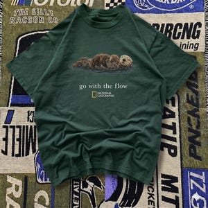 May include: Dark green t-shirt featuring a graphic of three otters floating in water. The text "go with the flow" and the National Geographic logo are printed below the otters. The shirt is made of a soft material.