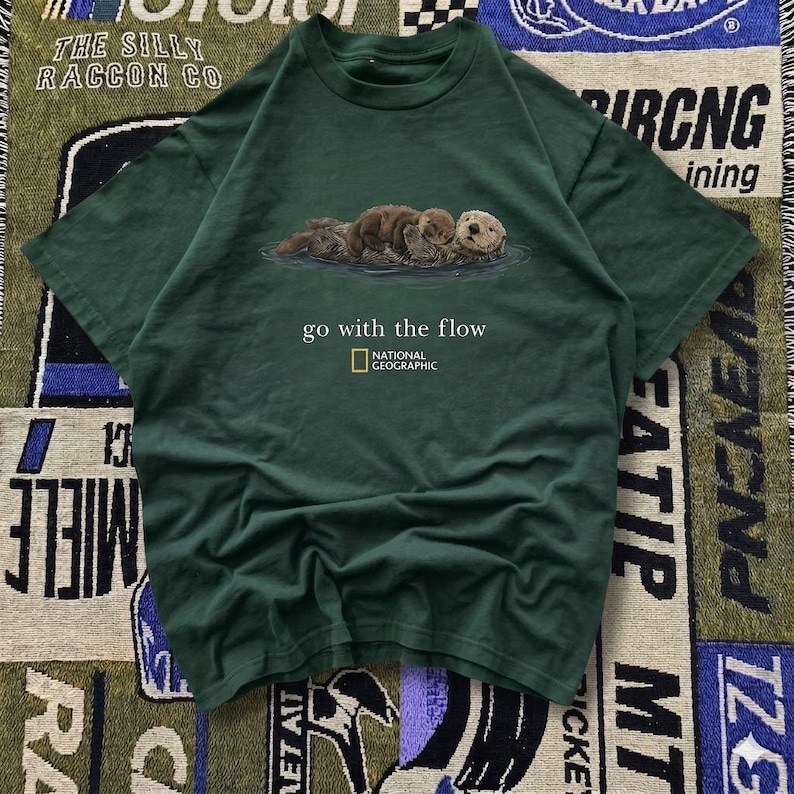 May include: Dark green t-shirt featuring a graphic of three otters floating in water. The text "go with the flow" and the National Geographic logo are printed below the otters. The shirt is made of a soft material.