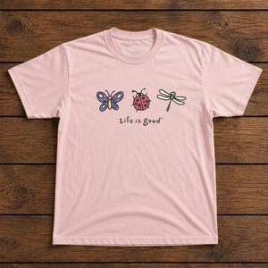 May include: A light pink t-shirt featuring a butterfly, ladybug, and dragonfly graphic. The text "Life is good" is printed below the illustrations. The shirt is made of a soft material.