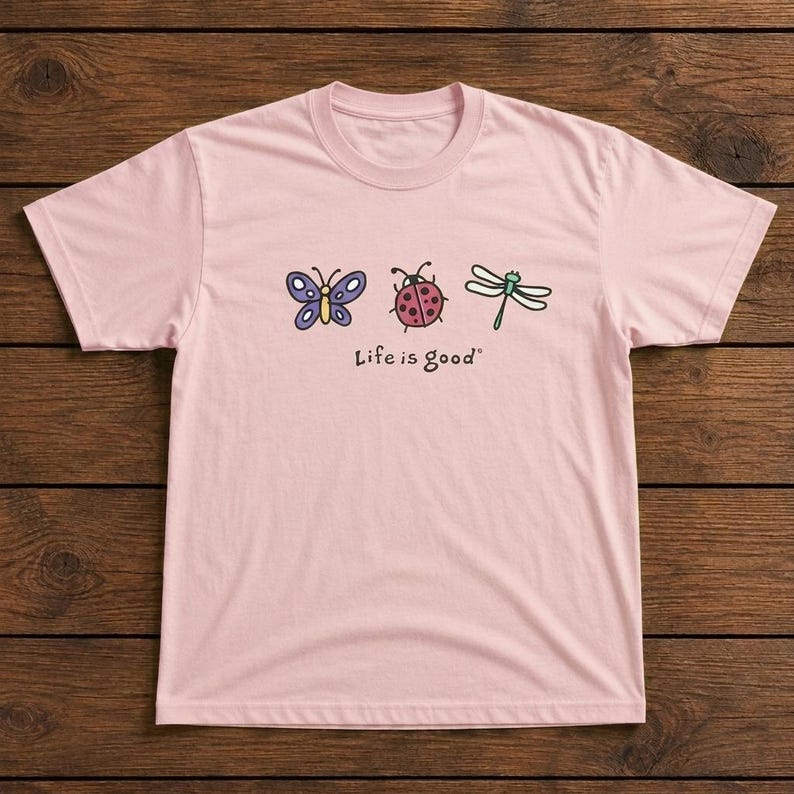May include: A light pink t-shirt featuring a butterfly, ladybug, and dragonfly graphic. The text "Life is good" is printed below the illustrations. The shirt is made of a soft material.