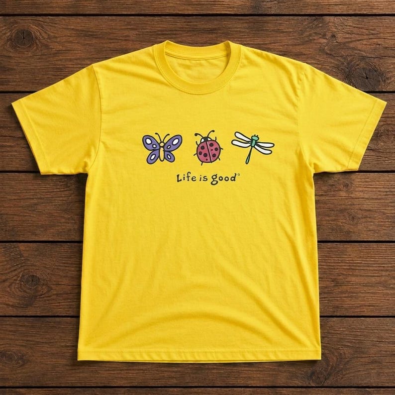 May include: A yellow t-shirt featuring a butterfly, ladybug, and dragonfly graphic. The phrase "Life is good" is printed below the illustrations. The shirt is made of a soft material and has a crew neck.