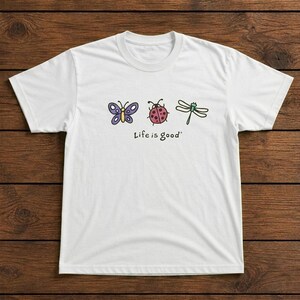 May include: White t-shirt featuring a colorful design of a butterfly, ladybug, and dragonfly. The phrase "Life is good" is printed below the illustrations. The shirt is made of a soft material.
