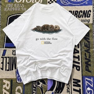 May include: White t-shirt featuring a graphic of three sea otters floating in water. The text "go with the flow" is printed below the image, with the National Geographic logo. The shirt is made of a soft material.