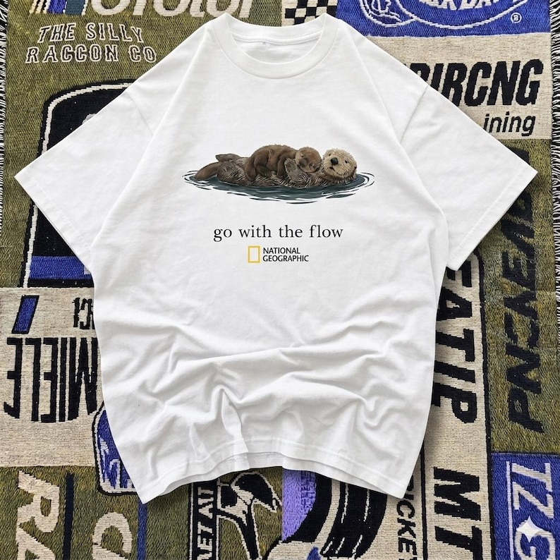 May include: White t-shirt featuring a graphic of three sea otters floating in water. The text "go with the flow" is printed below the image, with the National Geographic logo. The shirt is made of a soft material.