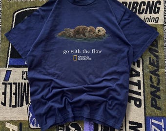 May include: Navy blue t-shirt featuring a graphic of sea otters floating in water. The text "go with the flow" and the National Geographic logo are printed below the otters. The shirt is made of a soft material.
