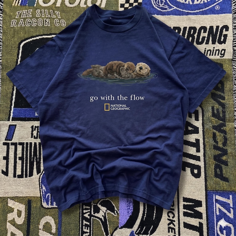 May include: Navy blue t-shirt featuring a graphic of sea otters floating in water. The text "go with the flow" and the National Geographic logo are printed below the otters. The shirt is made of a soft material.
