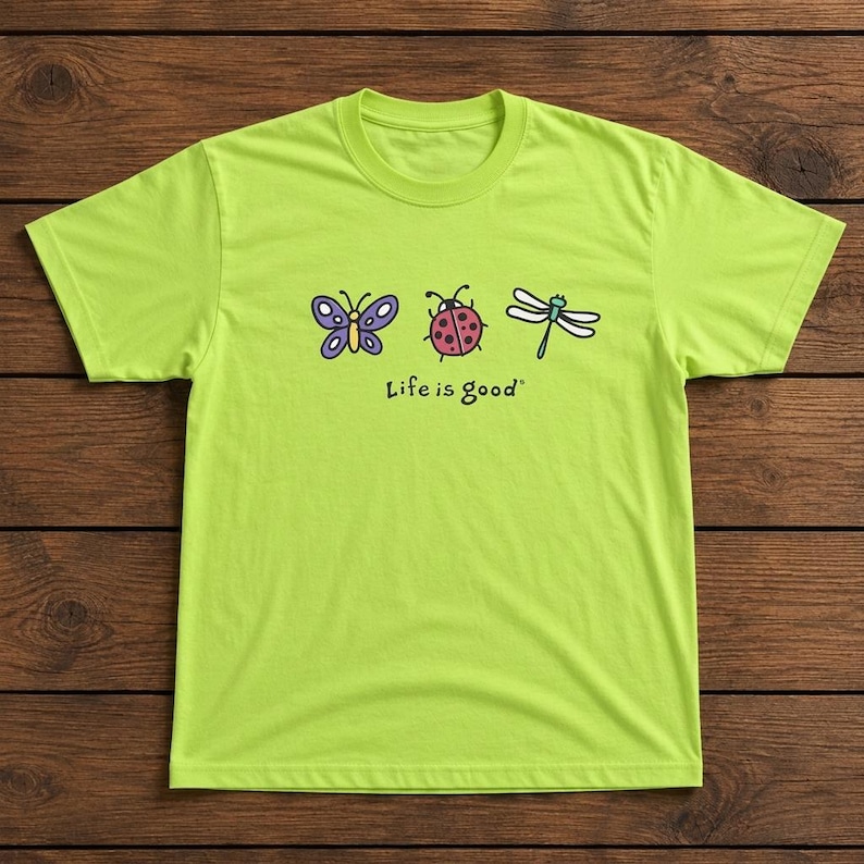 May include: A lime green t-shirt featuring a butterfly, ladybug, and dragonfly graphic. The butterfly is purple, the ladybug is red with black spots, and the dragonfly is white. The text "Life is good" is printed below the graphics.