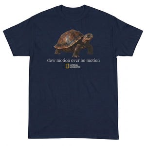 May include: Navy blue t-shirt featuring a tortoise with the text "slow motion over no motion" and the National Geographic logo. The tortoise is brown and has a textured shell. The shirt is a solid color and has short sleeves.
