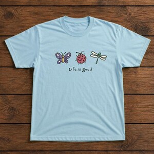 May include: Light blue t-shirt featuring a butterfly, ladybug, and dragonfly graphic. The phrase "Life is good" is printed below the illustrations. The shirt is made of a soft material and has a crew neck.