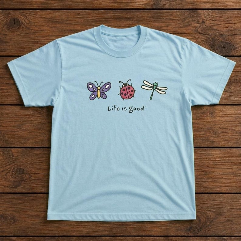 May include: Light blue t-shirt featuring a butterfly, ladybug, and dragonfly graphic. The phrase "Life is good" is printed below the illustrations. The shirt is made of a soft material and has a crew neck.
