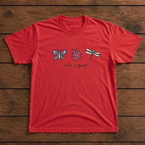 May include: A red t-shirt featuring a butterfly, ladybug, and dragonfly graphic. The phrase "Life is good" is printed below the illustrations. The shirt is made of a soft material and has a crew neck.