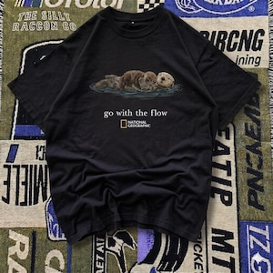 May include: Black t-shirt featuring an illustration of otters in water. The shirt has the text "go with the flow" and the National Geographic logo. The shirt is made of a soft material.