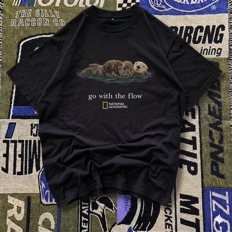 May include: Black t-shirt featuring an illustration of otters in water. The shirt has the text "go with the flow" and the National Geographic logo. The shirt is made of a soft material.
