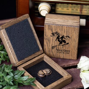 Könnte beinhalten: Eine quadratische Ringbox aus Holz mit Klappdeckel, die eine Lasergravur von zwei Personen beim Snowboarden auf einem Berg zeigt. Darunter ist der Text "NICK & GRACE" und "OCTOBER 26, 2026" eingraviert. Zwei Holzringe liegen auf schwarzem Schaumstoff.