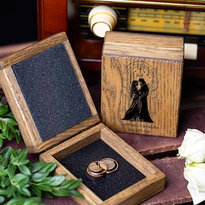 Puede incluir: Caja de anillos de madera con dos anillos de boda en su interior. La caja está abierta, revelando un interior negro y los anillos. La tapa de la caja presenta una ilustración negra de una pareja y el texto "Speak Friend and Enter".