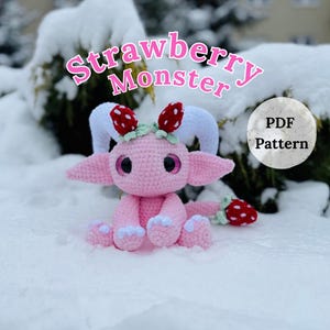May include: A pink crocheted Strawberry Monster doll with white horns and ears, and red strawberry accents. The doll is sitting in the snow. The image includes the text "Strawberry Monster" and "PDF Pattern".