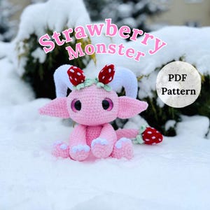 May include: A pink crocheted Strawberry Monster doll with white horns, large ears, and strawberry accents. The doll is sitting in the snow. The text "Strawberry Monster" is above the doll, and "PDF Pattern" is in a circle.