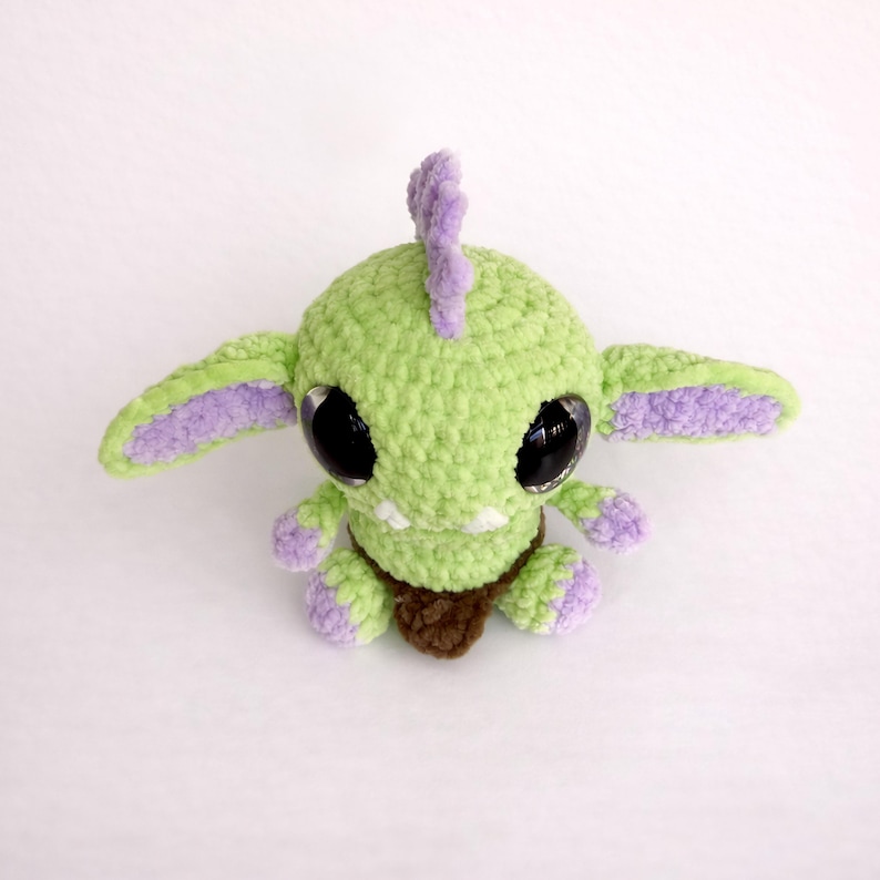 May include: A handmade crochet creature in shades of green, purple, and brown. The plush toy has large black eyes, a fin-like crest, and small limbs. The creature's body is a light green, with purple accents on the ears and feet.