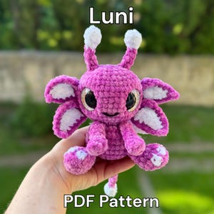 Könnte beinhalten: Ein handgefertigtes, plüschiges, magentafarbenes Wesen mit weißen Akzenten, großen schwarzen Augen und dem Text "Luni" und "PDF Pattern". Das Wesen hat mehrere blütenblattartige Anhänge und zwei Antennen mit weißen Pompons.