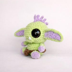 May include: A handmade crochet creature in shades of green and purple. The plush toy has large, glittery black eyes, pointed ears, and a small tuft of purple hair. It has small arms and legs with purple accents and a brown belt.