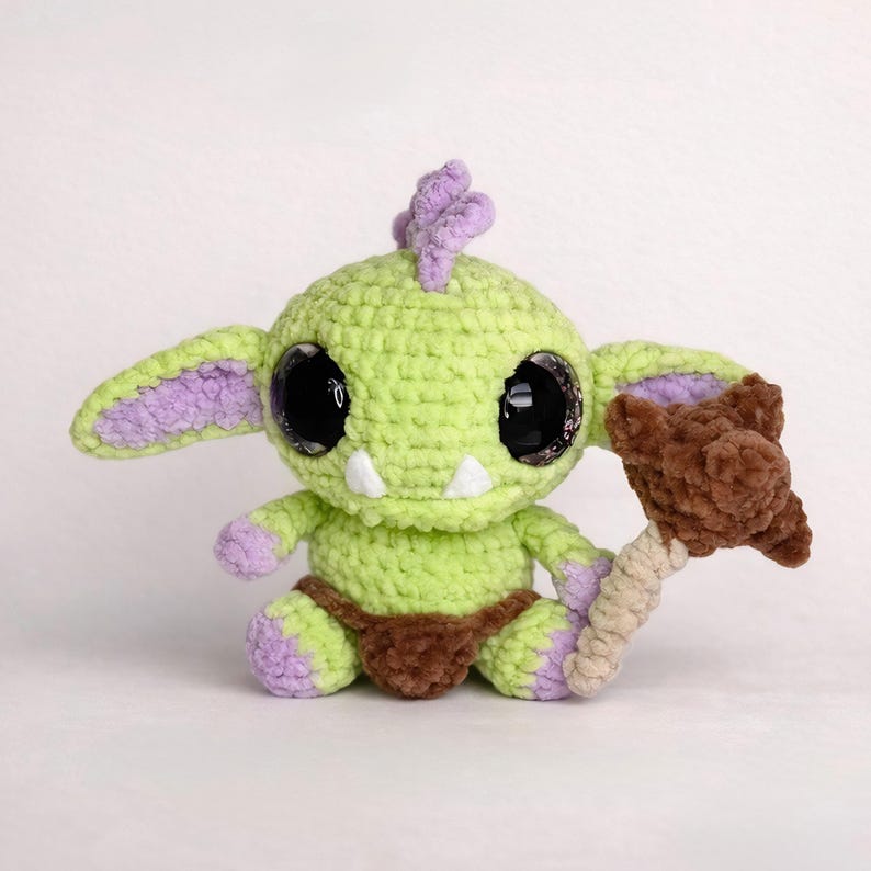 May include: A handmade crochet goblin doll in shades of green, purple, and brown. The doll has large, glittery black eyes, small white teeth, and a brown club. The goblin is sitting with its legs crossed.
