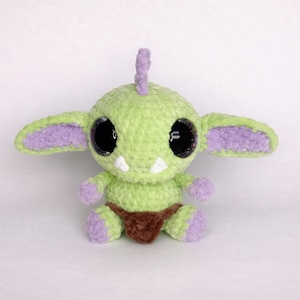 May include: A handmade crochet creature with a light green body, large black eyes, and purple accents on the ears and limbs. It has a small brown loincloth and two white teeth. The plush toy is a whimsical design.