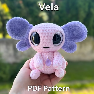 May include: A light pink crochet plush toy named Vela with large, dark, glittery eyes and purple, rounded, wing-like ears. The creature features a small, purple, teardrop-shaped pendant on its chest. The text Vela and PDF Pattern is visible.