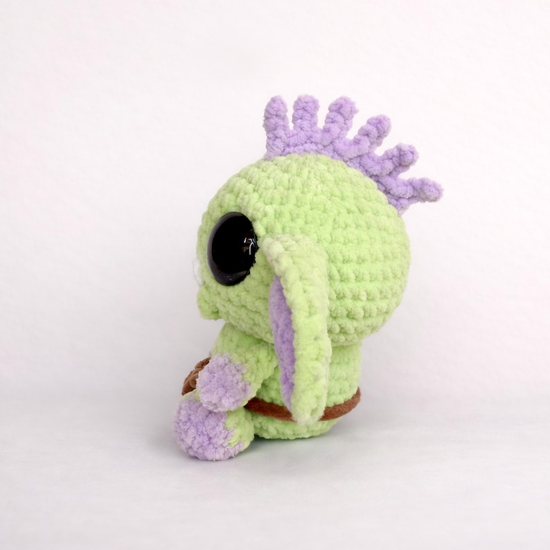 May include: A handmade, light green crocheted creature with large black eyes, purple accents, and a brown belt. The plush toy has a spiky, purple mohawk and purple-lined ears. The soft toy is sitting on a white surface.