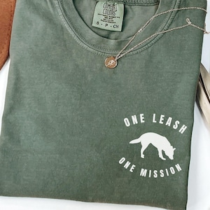 May include: An olive green short-sleeve t-shirt with a crew neckline. The shirt features a white graphic on the lower right side depicting a dog silhouette with the text ONE LEASH ONE MISSION. A thin gold-tone chain necklace rests on the collar.