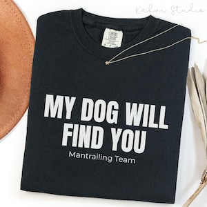 May include: A black crewneck t-shirt featuring the white text MY DOG WILL FIND YOU and Mantrailing Team printed on the front. The shirt has a Comfort Colors brand label at the inner neckline and is displayed with a thin gold-toned necklace.
