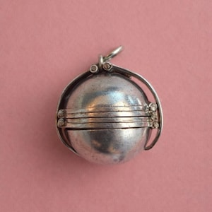 Può includere: Un pendente sferico color argento con un design a cerniera. Il pendente ha un anello nella parte superiore per una catena ed è composto da più fasce orizzontali. Lo sfondo è di colore rosa.