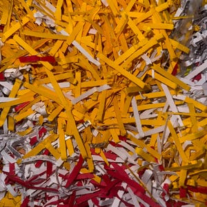 Pode incluir: Uma visão de perto de papel picado em amarelo, vermelho e branco. As tiras de papel têm comprimentos e larguras variados, criando um padrão texturizado e abstrato. As cores são vibrantes e o efeito geral é caótico.