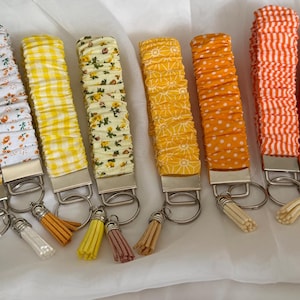 Puede incluir: Una colección de llaveros de tela con varios estampados y colores. Los diseños incluyen flores, cuadros y lunares en amarillo, naranja y blanco. Cada llavero tiene un cierre de metal plateado y una borla a juego.