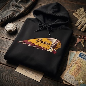 May include: Black hoodie with the "Indian Motorcycle" logo in yellow, maroon, and white. The design features a Native American profile and the words "Indian Motorcycle". The hoodie is on a wooden surface with other accessories.